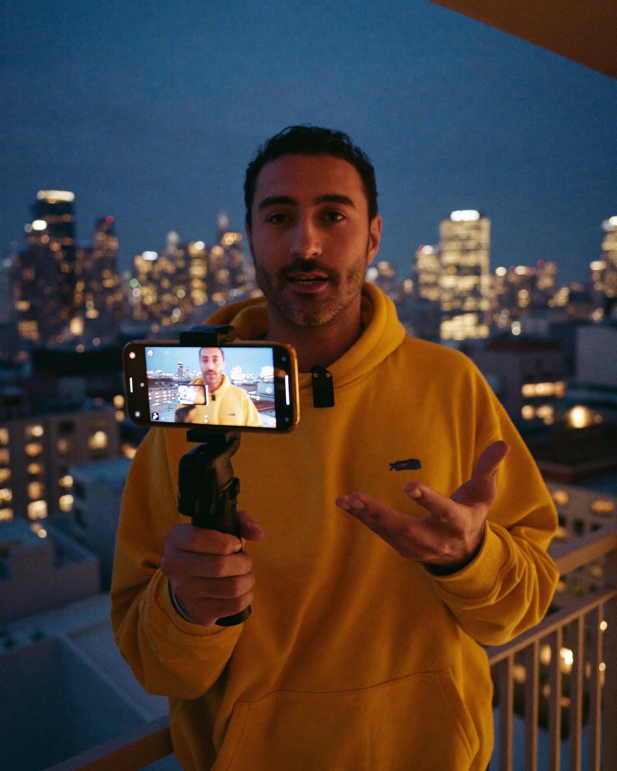 Creator na varanda à noite, gravando a si mesmo com o celular num estabilizador